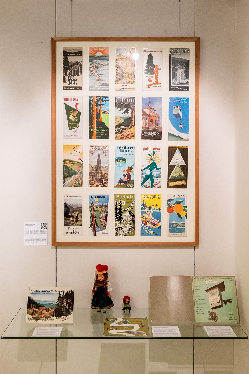 20 diverse Prospekte in einem großflächigen Holzrahmen an der Wand. Auf einem Regal darunter befindet sich ein aufgeklapptes Buch, ein Spiel, zwei Puppen in Schwarzwälder Tracht und eine Schwarzwald-Postkarte.