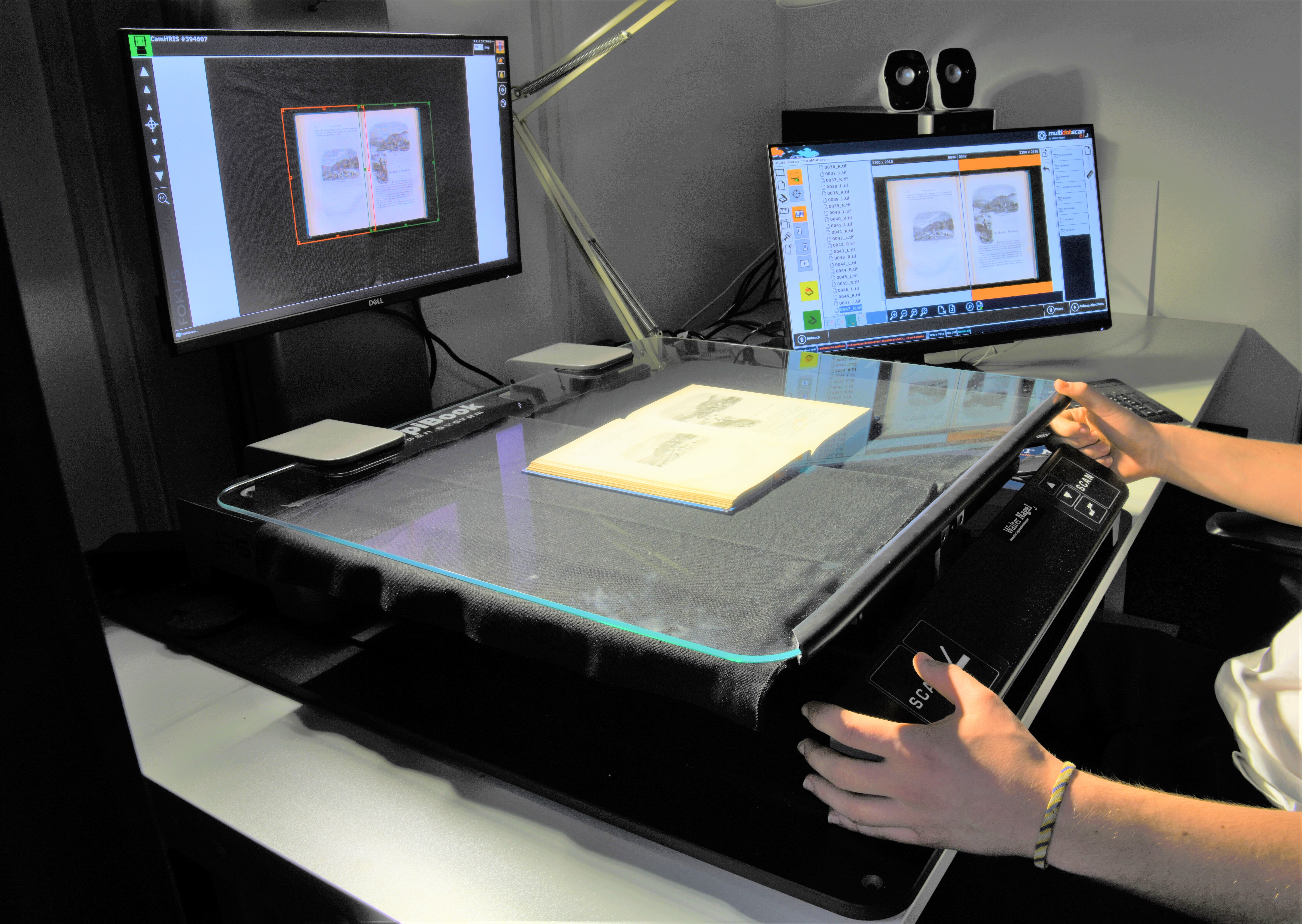 Das Bild zeigt einen Copibook-Scanner auf einem Arbeitstisch. Ein Buch liegt aufgeklappt auf der Oberfläche des Geräts. Die Hände einer Mitarbeiterin liegen entspannt seitlich am Copibook.