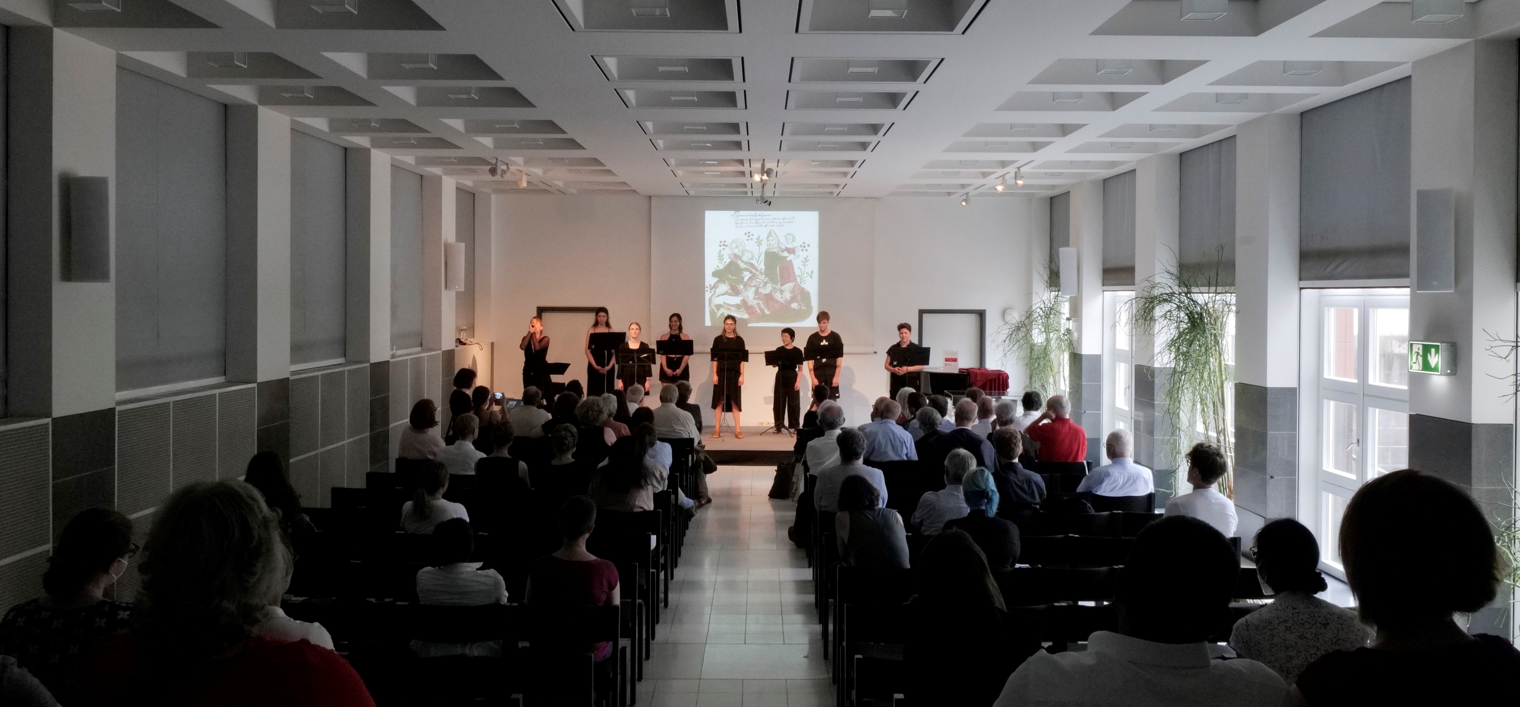 Aus dem Hintergrund des Saals blickt man über das Publikum nach vorn auf die Bühne, wo die sieben in Schwarz gekleideten Studierenden den Text des Wigalois in Mittelhochdeutsch vortragen.