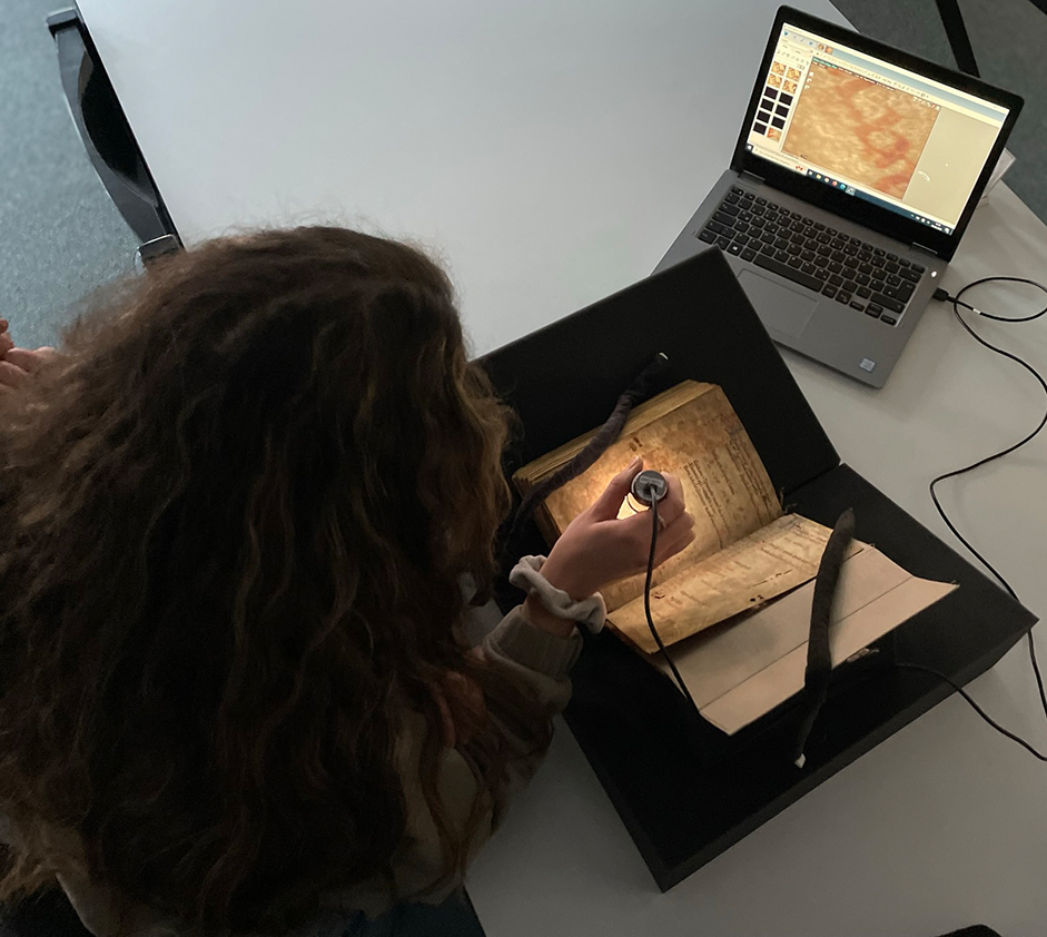 Zu sehen ist das Digitalmikroskop, das von einer Nutzerin bei einem alten gedruckten Buch eingesetzt wird.
