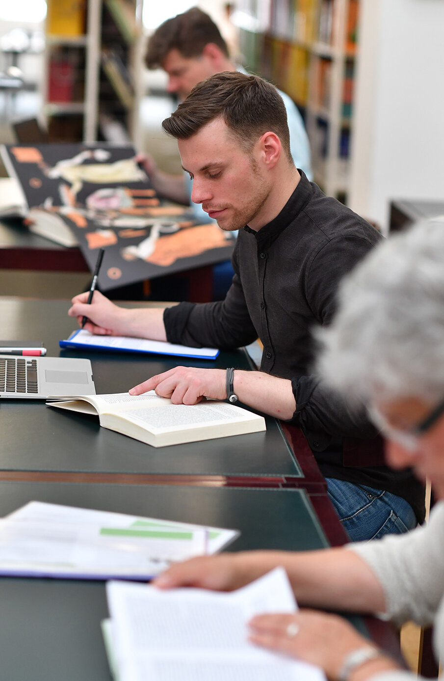 Zu sehen sind drei Personen. Allesamt sitzen diese an Tischen und studieren Bücher. Die Person sind linear aufgereiht, der Bildfokus liegt auf der mittleren Person. Die beiden flankierenden Personen sind mit einer Unschärfe überlagert.  