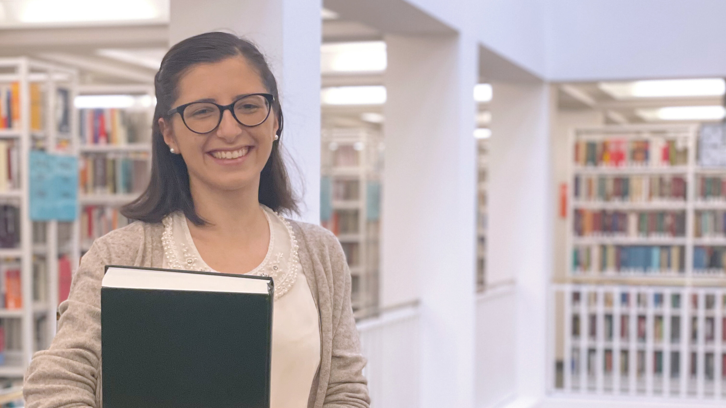 © Badische Landesbibliothek Zu sehen ist das erste Obergeschoss der BLB. Im Vordergrund steht auf der linken Bildseite eine junge Frau. Sie lächelt freundlich und hat ein Buch in den Armen. Im Hintergrund sind zahlreiche Bücherregale zu sehen.