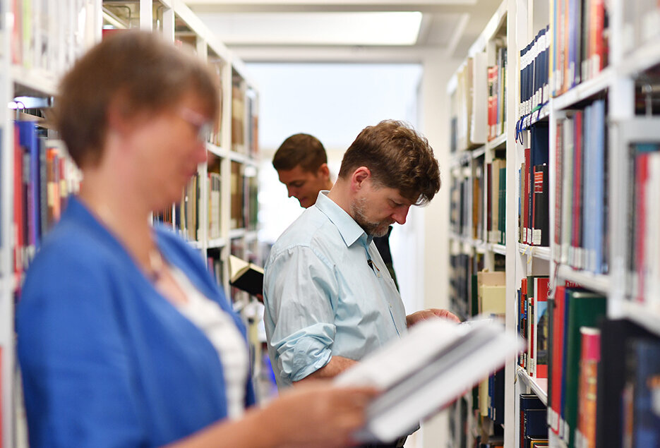 Zu sehen sind mehrere Personen. Diese stehen zwischen den Bücherregalen BLB. Im Vordergrund steht eine Frau mit einem aufgeschlagenen Buch in der Hand. 