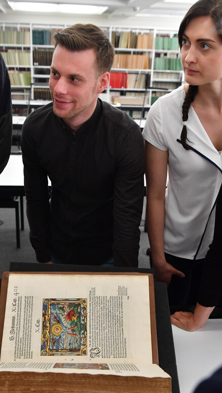 Das Bild zeigt einen Mann und eine Frau, die im Rahmen einer Einführung in die historischen Bestände im Lesesaal Sammlungen der Badischen Landesbibliothek gespannt einer Mitarbeiterin zuhören. Vor ihnen auf dem Tisch liegt eine Inkunabel, ein Wiegendruck. 