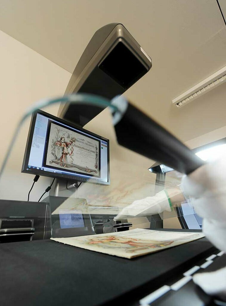 An einem Buchscanner der Digitalisierungswerkstat in der Badischen Landesbibliothek wird ein querformatiges Heft digitalisiert.