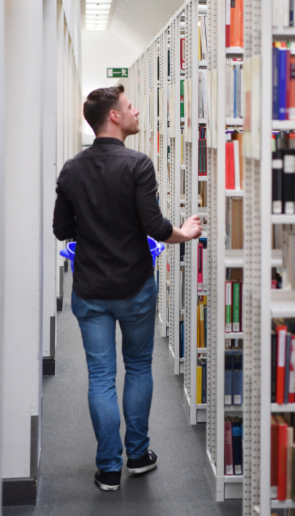 Das Bild zeigt in Rückenansicht einen Nutzer, der mit einem Bücherkorb durch die Gänge des offenen Magazins läuft. 