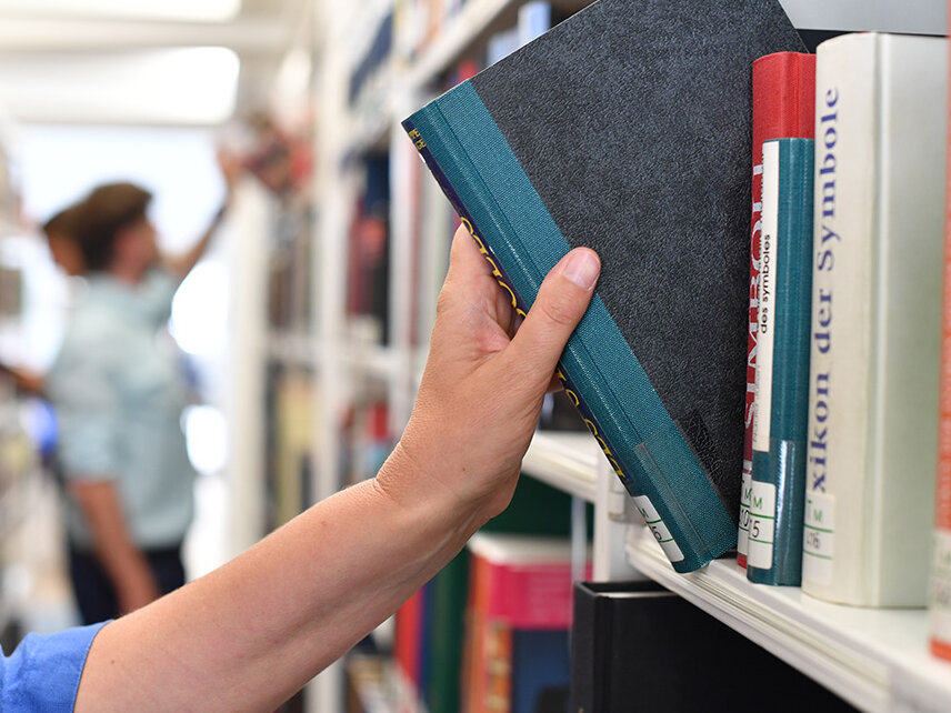 Eine Hand zieht gerade ein Buch aus einem Bücherregal. Im Hintergrund ist eine Person zu sehen, die ebenfalls ein Buch aus dem Bücherregal entnimmt. 