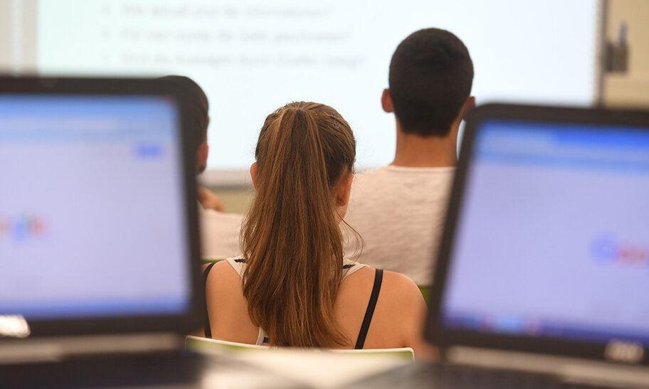 © Foto: Uli Deck Zu sehen sind im Vordergrund zwei Notebooks. Diese nehmen den rechten und den linken Bereich des Bildes ein. Die Lücke zwischen beiden gibt Blick auf Personen in einer Schulungssituation frei. Diese sind von hinten abgelichtet worden und unscharf.