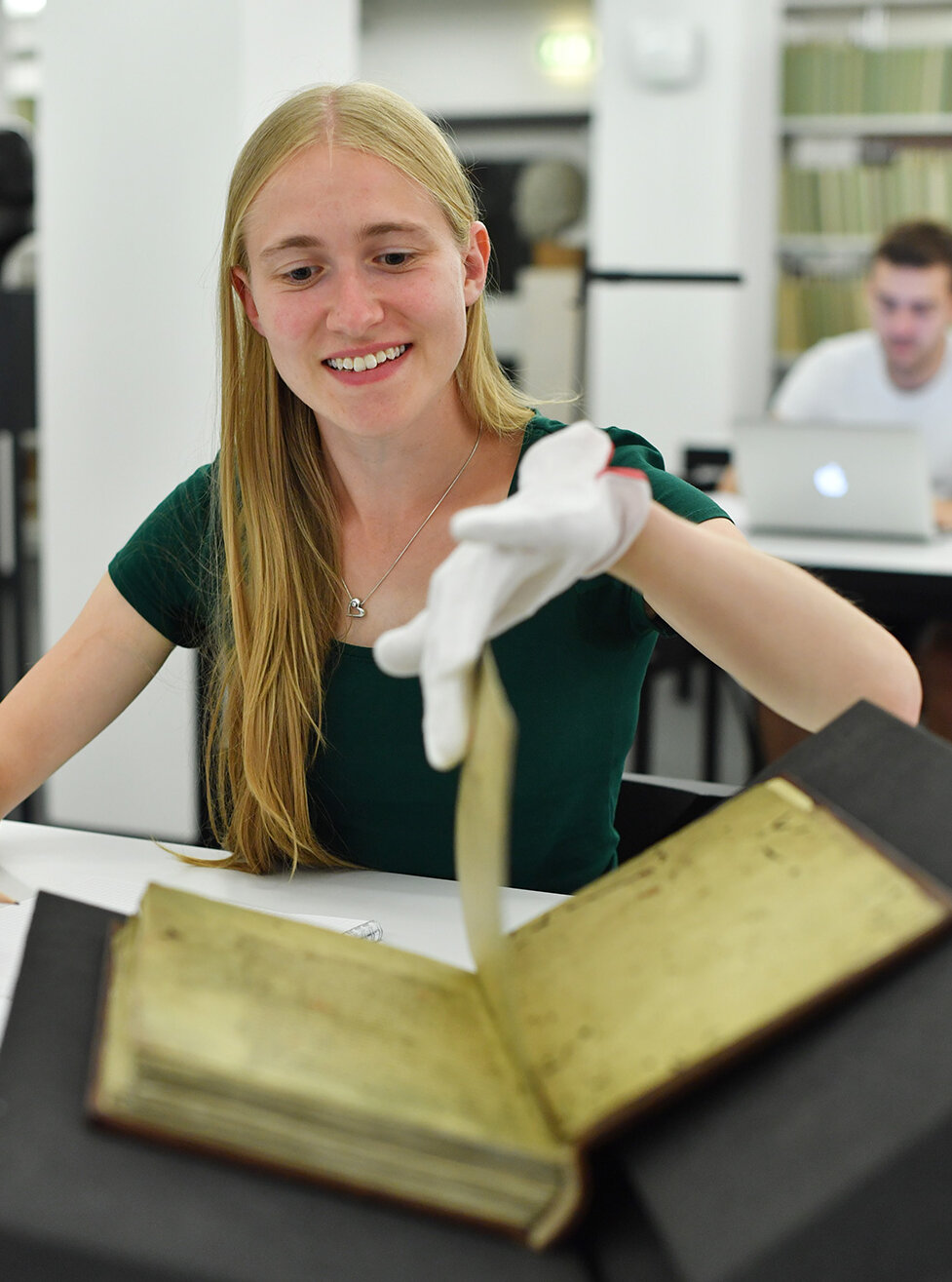 Zu sehen ist eine lächelnde junge Frau mit weißen Handschuhen, die grade eine Seite in einem historischen Buch umblättert. 