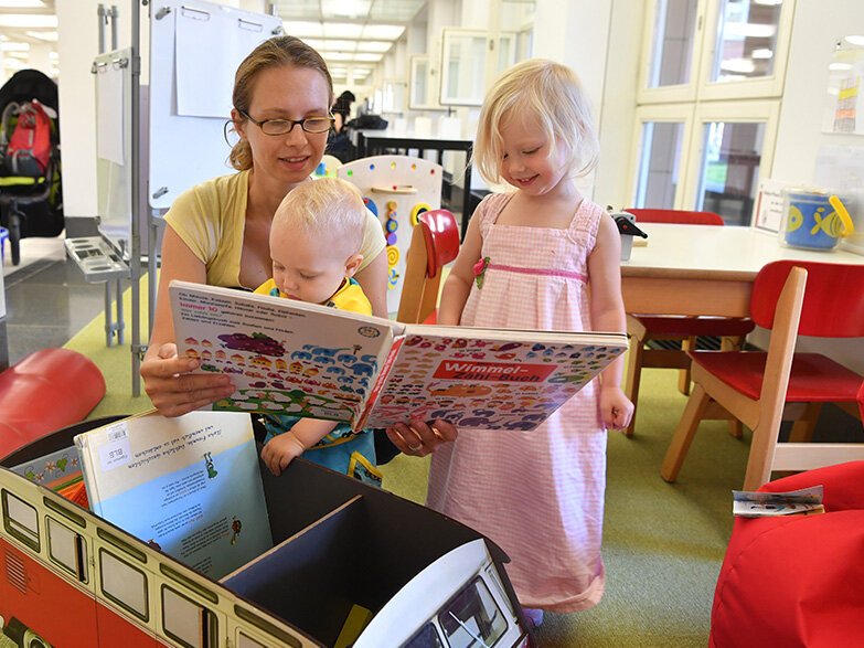 Auf dem Bild sind drei Personen zu sehen. Zwei Kleinkinder und eine Erwachsene. Sie schauen zusammen ein Buch in der Spielecke der BLB an. 