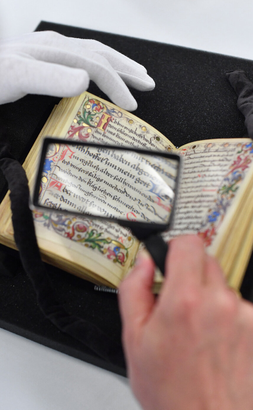 Das Bild zeigt einen Bibliotheksnutzer, der sich mit Handschuh und Lupe ein aufgeschlagenes Werk aus dem historischen Sammlungsbestand der Badischen Landesbibliothek näher ansieht.