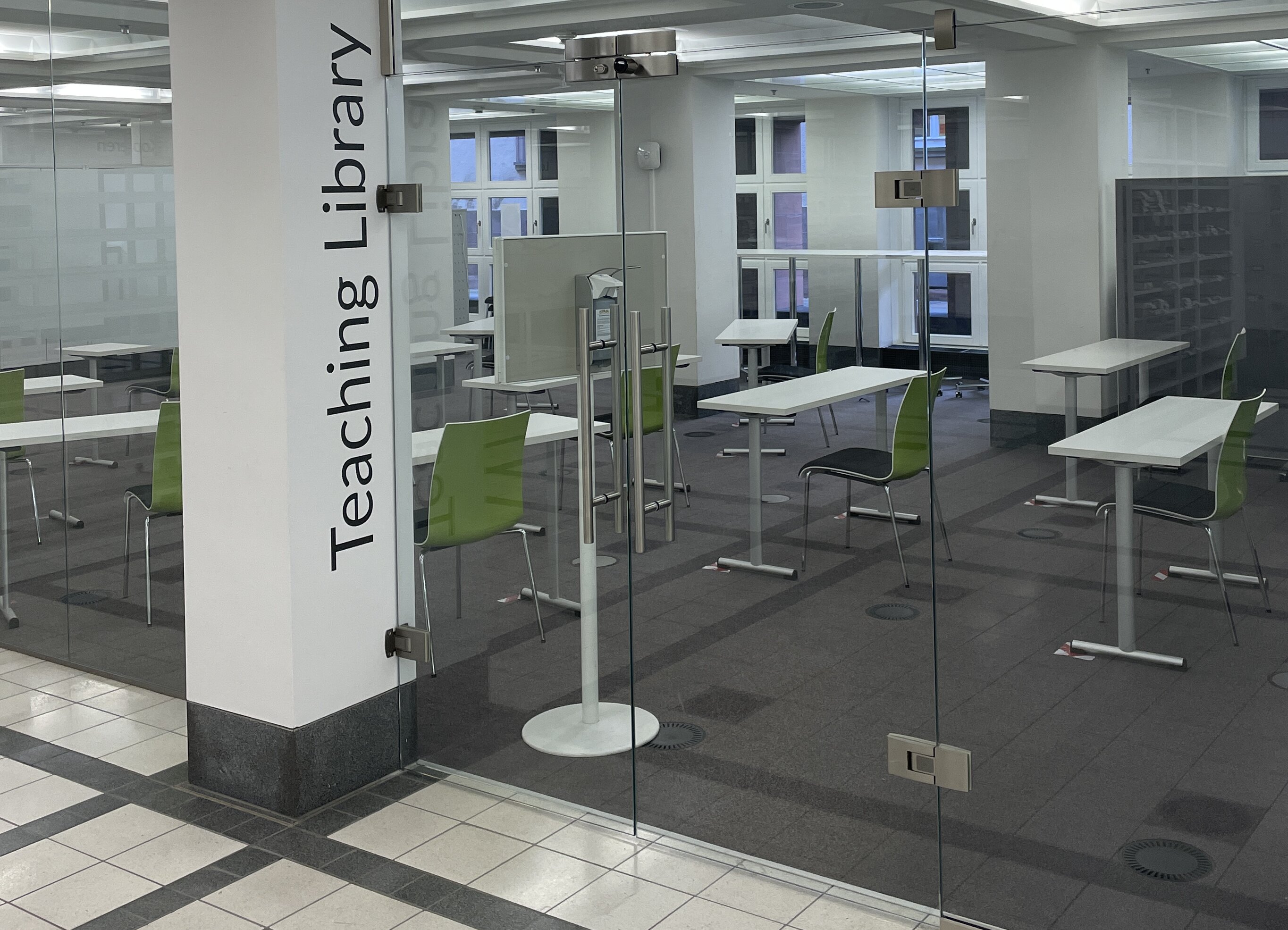 Schulungsraum der Teaching Library Zu sehen ist der Eingangsbereich des Schulungsraums der Teaching Library im Foyerd er BLB. Die Eingangstür ist komplett aus Glas und gibt Blick auf den dahinterliegenden Schulungsraum frei. Dort stehen mehre helle Tische und grüne Stühle. neben der Eingangstür ist in schwarzen Buchstaben auf weißer Wand Teaching Library zu lesen.