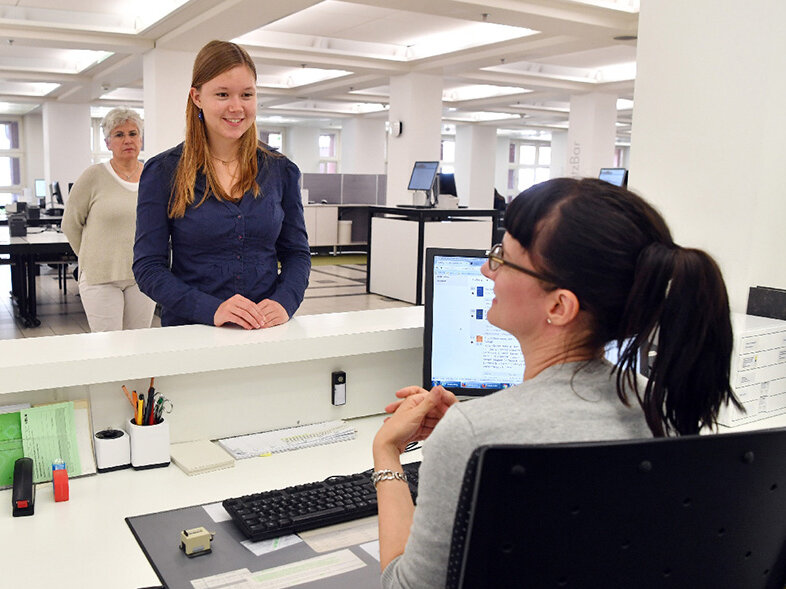 Das Bild zeigt eine Mitarbeiterin der Badischen Landesbibliothek, die einer Nutzerin am Infoschalter Auskunft gibt. Im Hintergrund steht eine weitere Nutzerin in der Warteschlange.