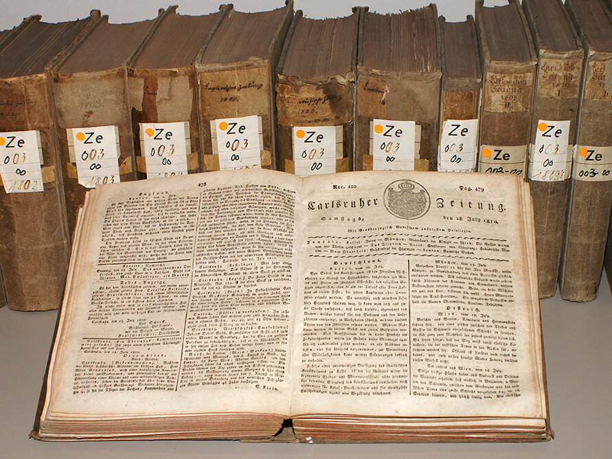 Im Vordergrund ist ein offenes Buch, die aufgeschlagene Seite lässt eine Titelseite der "Karlsruher Zeitung erkennen. Im Hintergrund steht eine Reihe in Bände gebundener Zeitung.