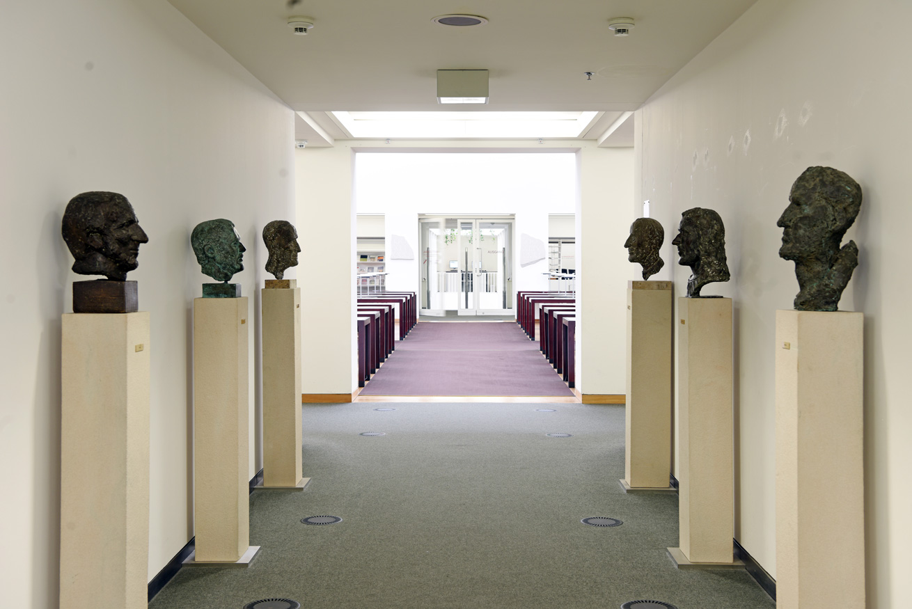 Das Bild zeigt sechs Bronzebüsten des Künstlers Werner Stötzer, die entlang des zentralen Durchgangs zwischen dem Kuppelbau und dem hinteren Bereich des Lesesaals aufgestellt sind.