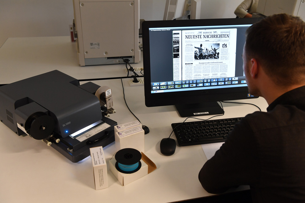 Das Bild zeigt einen Mann in Rückenansicht, der an einem der Mikrofilm-Arbeitsplätze im Lesesaal der Badischen Landesbibliothek sitzt. Auf dem Bildschirm des PCs ist das Titelblatt einer Ausgabe der Badischen Neuesten Nachrichten zu sehen.