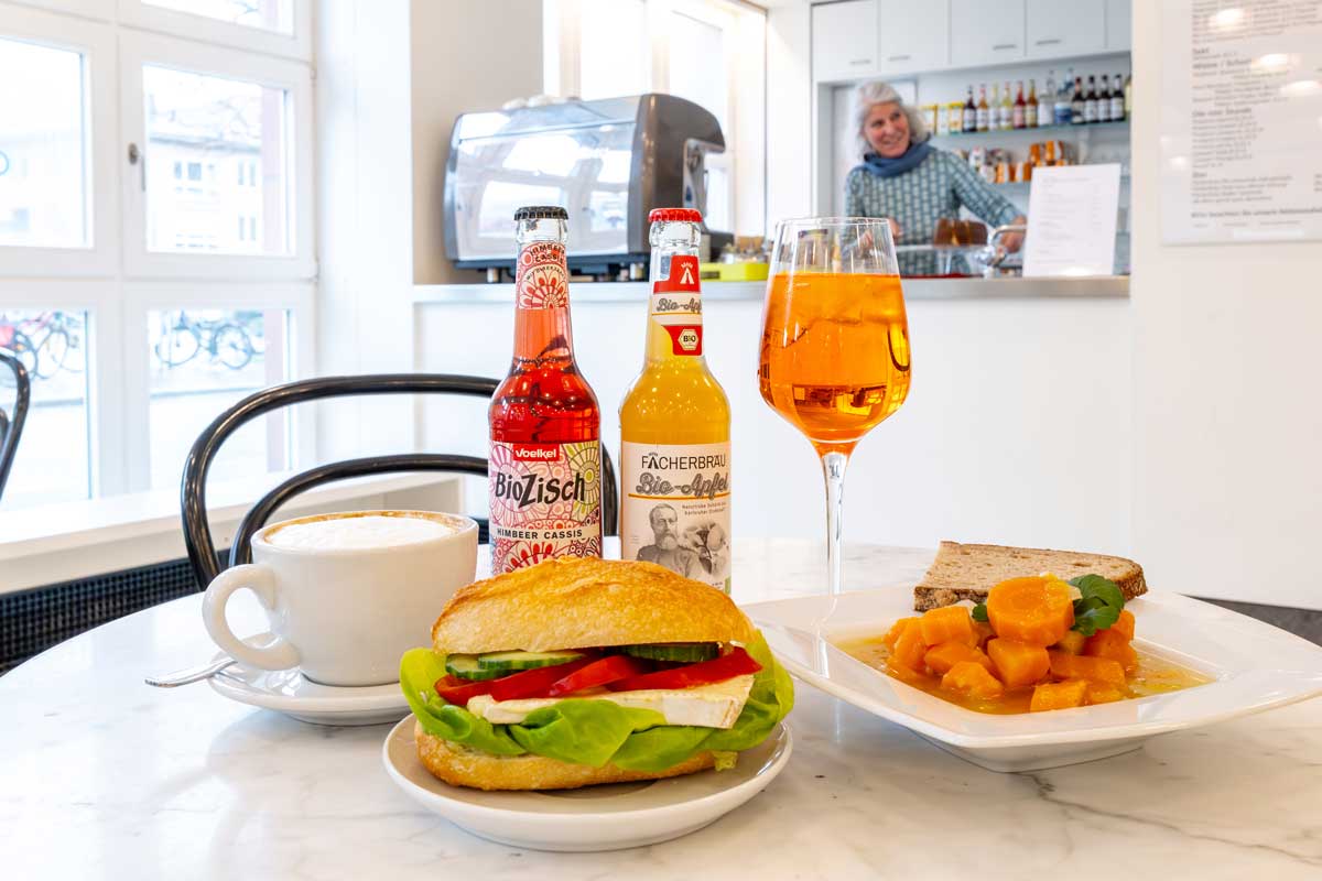 Das Foto zeigt den Innenbereich des Cafés, im Vordergrund eine Auswahl an Speisen und Getränken.