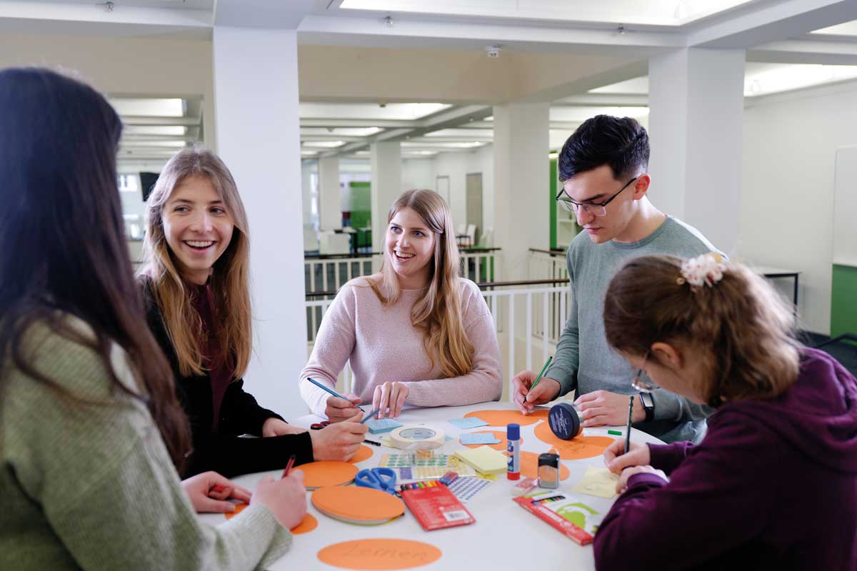 Zu sehen sind mehrere Personen. Sie sitzen an einem Tisch mit verschiedenen Lernmaterialien. Sie unterhalten sich und arbeiten mit den Materialien.