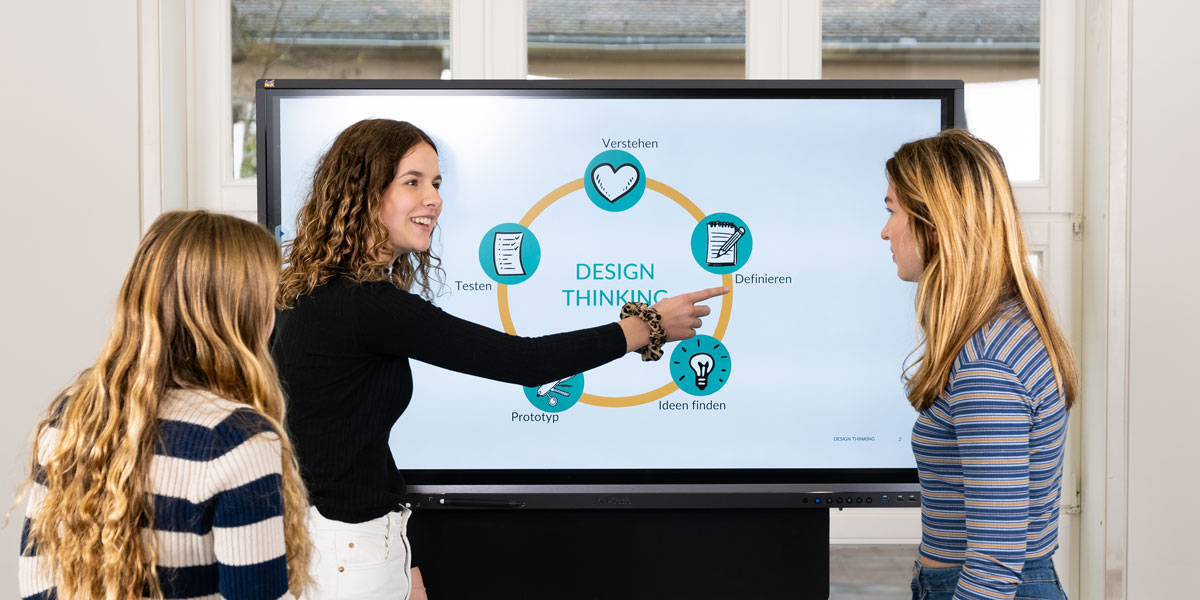 Drei junge Frauen stehen an einem Bildschirm und unterhalten sich über die gezeigte Abbildung. Diese visualisiert den Kreislauf der Methode „Design Thinking“.