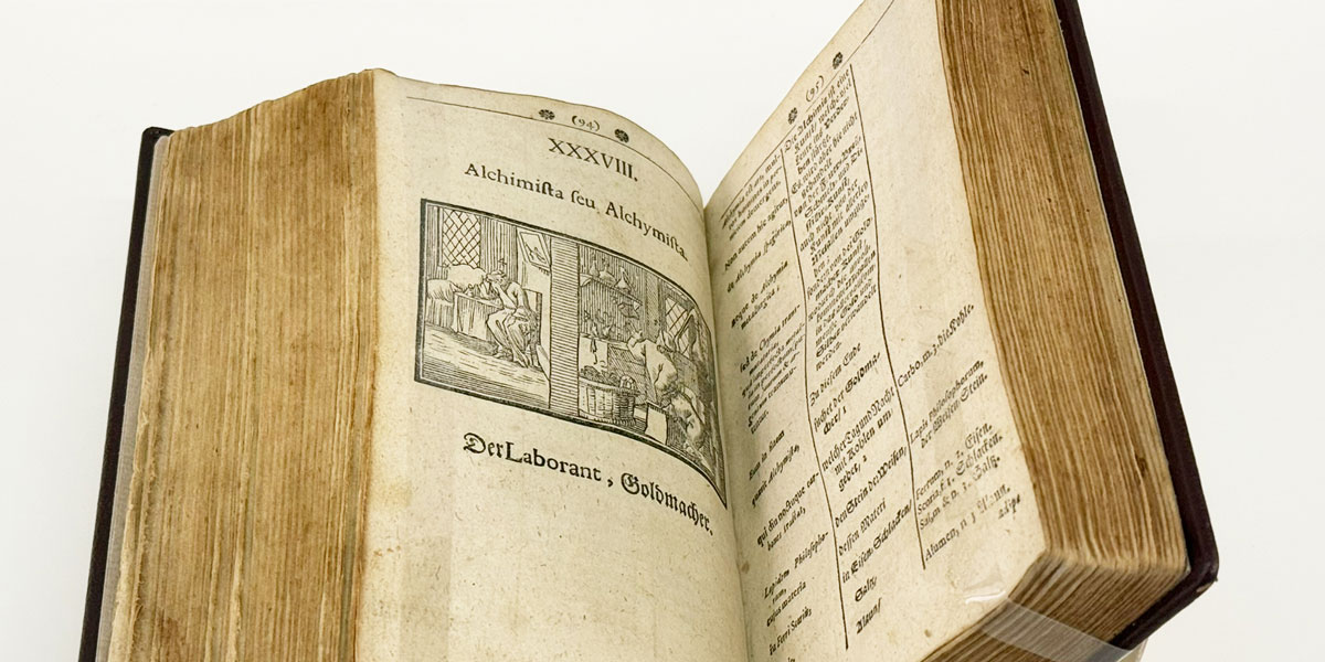 Zu sehen ist ein aufgeschlagenes Buch. Auf der linken Seite ist eine Illustration mit dem Untertitel „Der Laborant, Goldmacher.“ abgebildet.