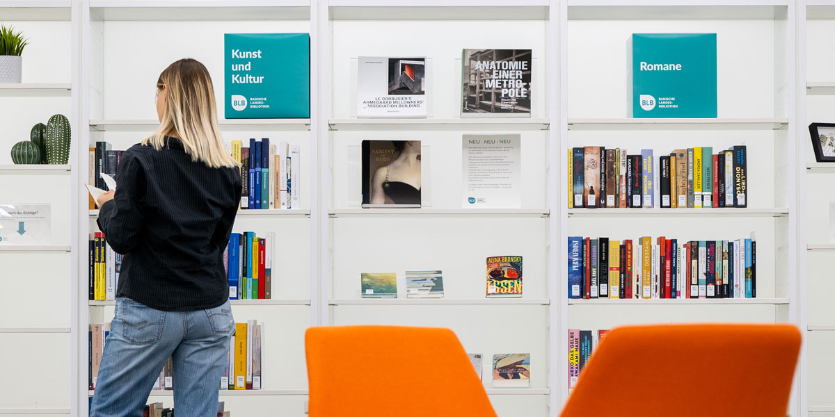 Eine Frau steht vor dem Neuerwerbungsregal und schaut sich ein Buch an. Im Vordergrund sind zwei orangefarbene Sessel zu erahnen.