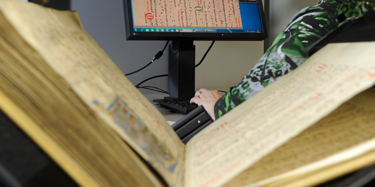 Zu sehen ist ein aufgeschlagenes Buch. Im Hintergrund ist ein Bildschirm angeschnitten zu erkennen und eine Person bedient das Programm.