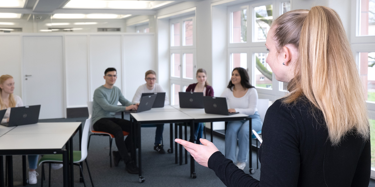 Seminarraum mit einer Gruppe Studierender an Tischen mit Laptops und einer Dozentin, die ihnen zugewandt steht und spricht.