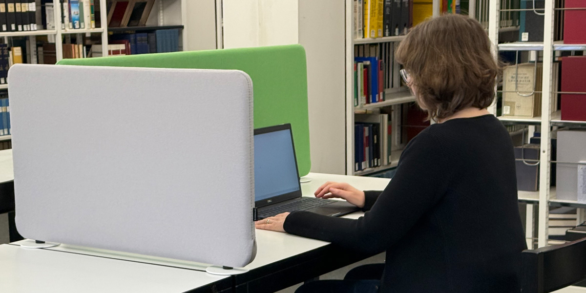 Person sitzt an einem Arbeitsplatz im Lesesaal, der durch einen Sichtschutz abgetrennt ist, und arbeitet an einem Laptop.