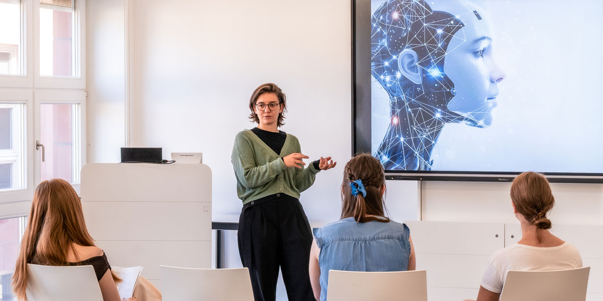 Referentin steht vor Gruppe von drei Personen in Seminarraum und hält Präsentation mit digitaler Grafik eines menschlichen Kopfes mit Netzwerkstruktur auf Bildschirm.