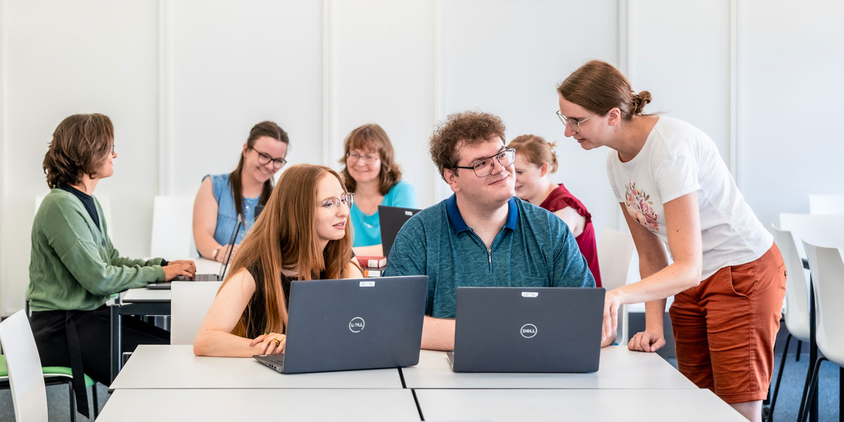 Gruppe von Erwachsenen sitzt an Tischen mit Laptops in einem hellen Seminarraum, eine Frau steht und spricht mit einem Mann und einer Frau, die an einem Laptop arbeiten.