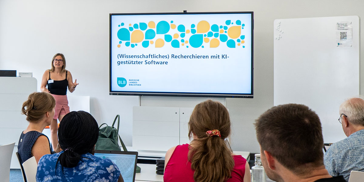 Frau steht vor großem Bildschirm mit Präsentation zum Thema wissenschaftliches Recherchieren mit KI-gestützter Software, mehrere Personen sitzen mit Laptops im Seminarraum der Lernwerkstatt.
