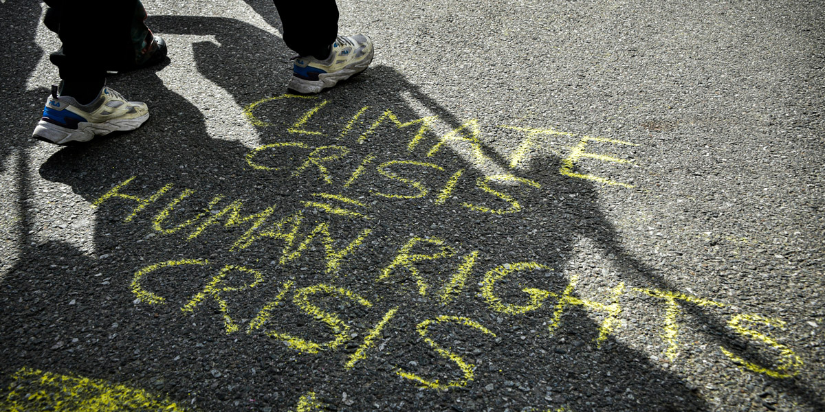 Auf dem Asphalt einer Straße ist „Climate Crisis = Human Rights Crisis“ mit gelber Kreide geschrieben. Daneben sind die Beine von Personen zu sehen, die marschieren.