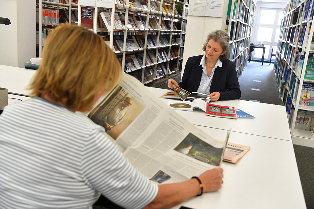 Das Bild zeigt die Rückenansicht einer Bibliotheksnutzerin, die an einem Tisch sitzend in einer Zeitung liest. Ihr gegenüber sitzt eine weitere Bibliotheksnutzerin, die in einem Magazin liest.