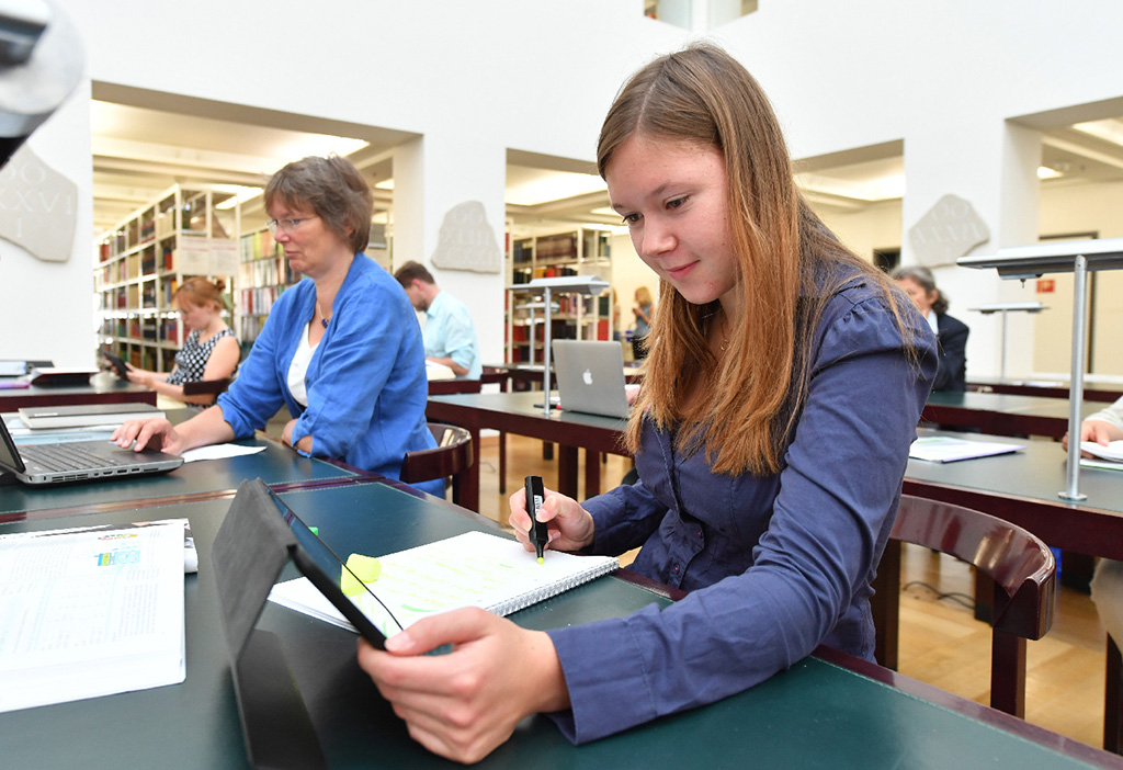 Das Bild zeigt Nutzerinnen und Nutzer an den Arbeitsplätzen im Lesesaal der Badischen Landesbibliothek. Vorne im Bild ist eine Bibliotheksnutzerin damit beschäftigt, etwas farbig in ihren Notizen zu markieren, während sie gleichzeitig auf den Bildschirm eines Tablets blickt.