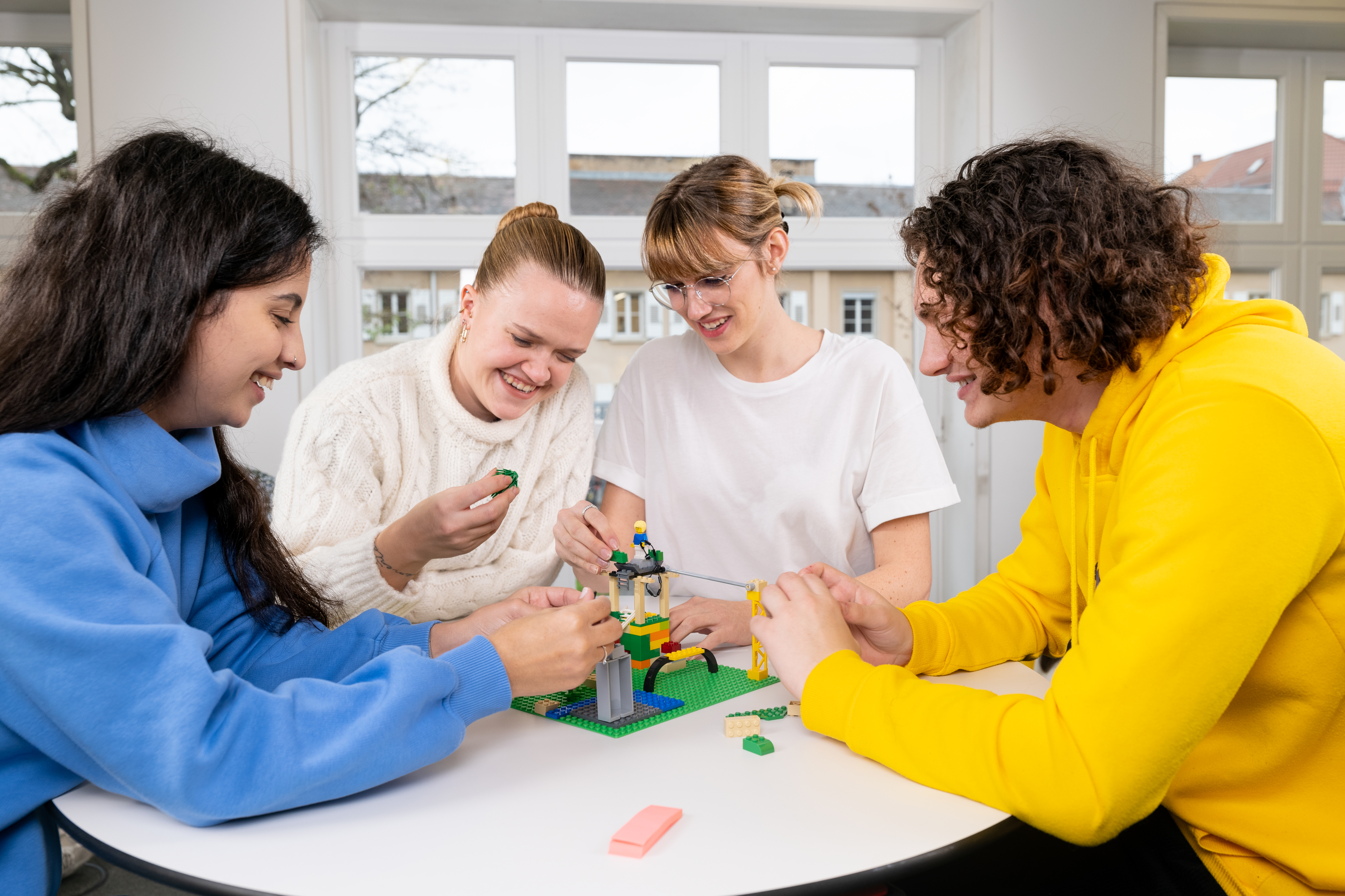 Zu sehen sind vier junge Personen, die an einem Tisch sitzen und mithilfe von LEGO©-Steinen ihre Ideen visualisieren.