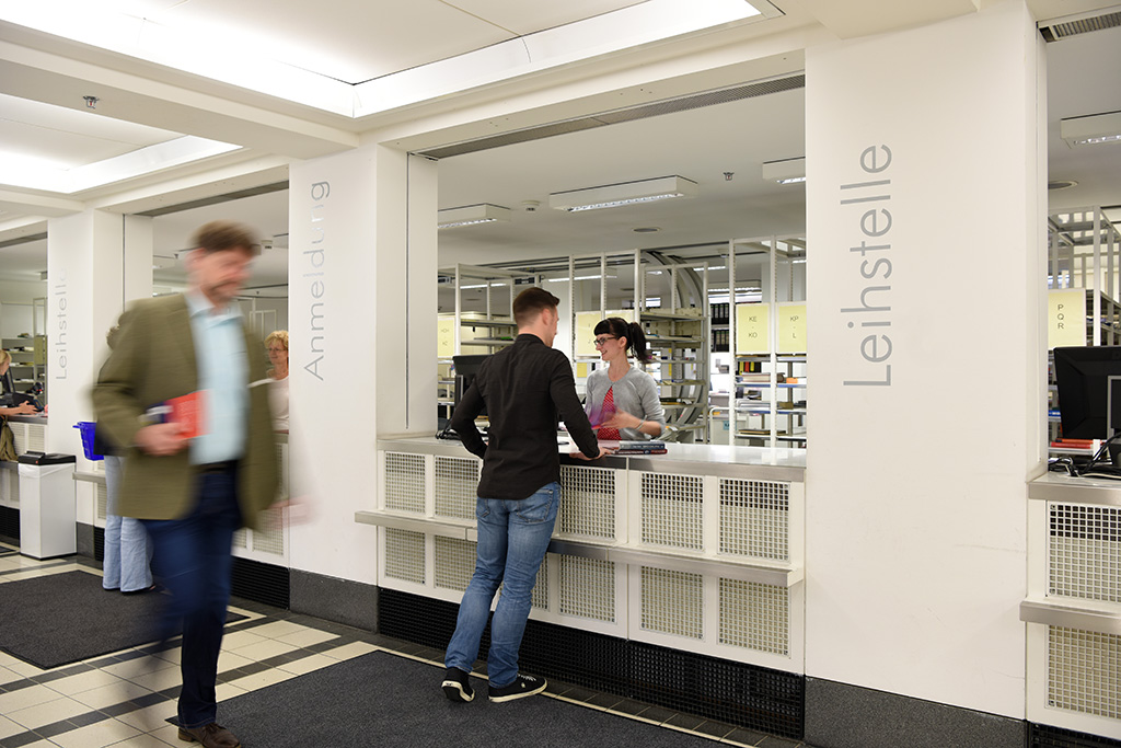 Langzeitbelichtung des Anmeldeschalter. Ein Besucher meldet sich bei einer Mitarbeiterin am Schalter an, im Vordergrund läuft ein Mann mit ausgeliehenen Büchern vorbei.