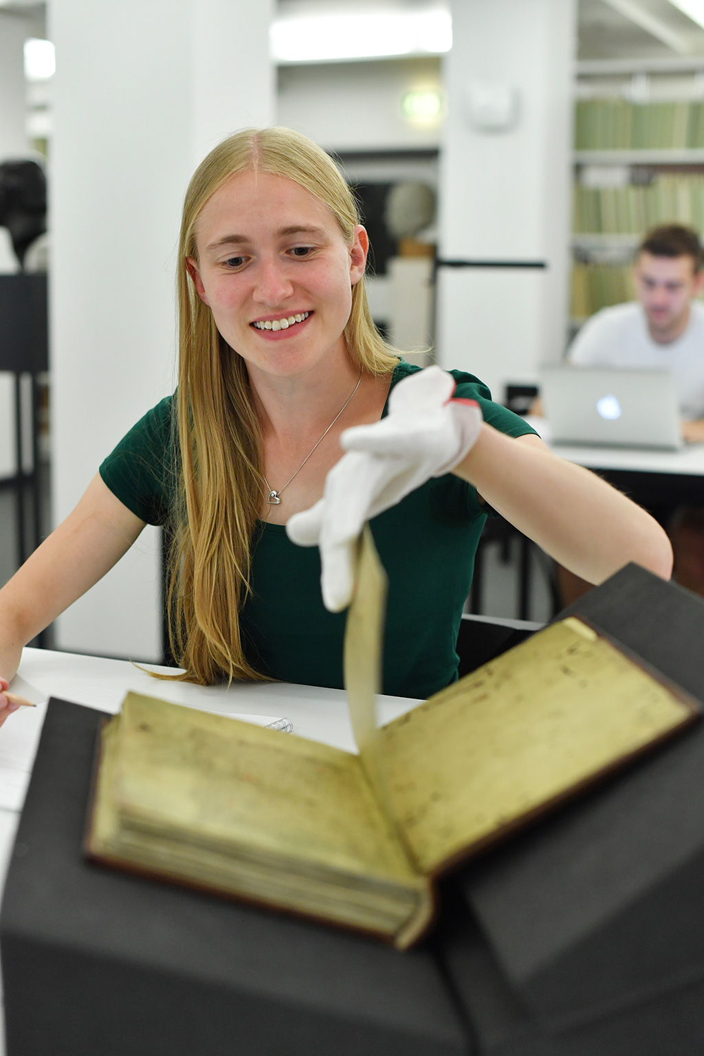 Zu sehen ist ein aufgeschlagenes Buch im Lesesaal Sammlungen. Eine junge Frau blättert in diesem historischen Buch gerade eine Seite um. Sie lächelt und trägt weiße Handschuhe.  