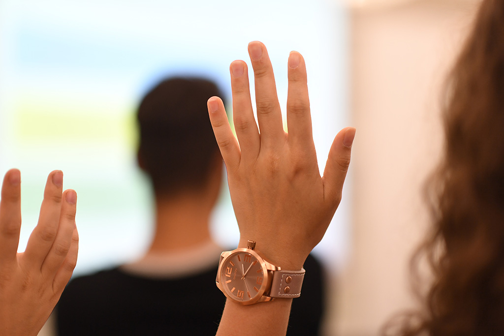 Zu sehen sind zwei Personen. Diese wurden von hinten fotografiert und heben jeweils eine Hand um sich zu melden. Der Bildausschnitt zeigt nur die Köpfe und die gehobenen Hände. 