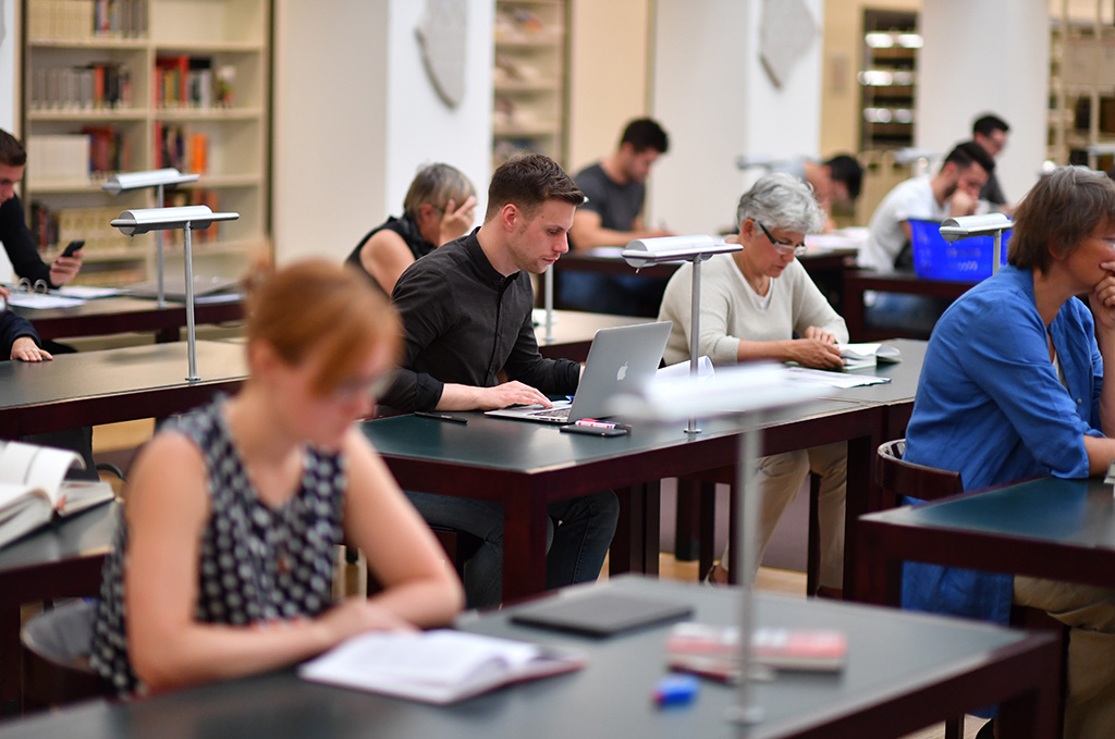 Das Foto zeigt den Lesesaal der BLB. An den Tischen sitzen mehrere Personen und arbeiten mit Büchern und Notebooks. Die Bildschärfe konzentriert sich dabei auf zwei Personen in der Bildmitte. 