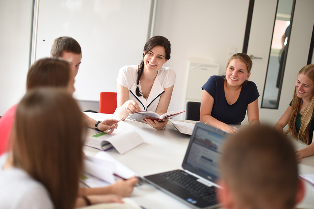 Zu sehen ist eine Gruppe von Personen. Diese gruppiert sich um einen Tisch und befindet sich im Austausch. Auf dem Tisch sind Bücher und ein Notebook zu sehen.  