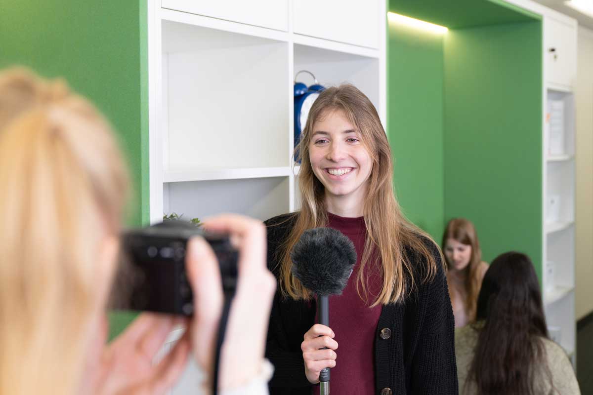 Zu sehen sind ist eine Person in Rückansicht mit einer Kamera in der Hand. Ihr gegenüber steht eine lächelnde Person mit einem Mikrophon in der Hand. Im Hintergrund sind weitere Personen zu erkennen. 