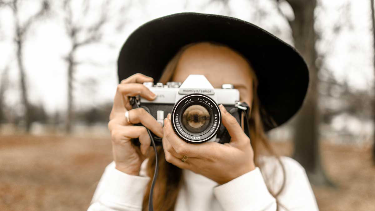 Zu sehen ist eine Fotografin. Sie fotografiert direkt in Richtung des Betrachters. Die kamera verdeckt Ihr Gesicht, sie trägt einen Hut und einen weißen Pullover. Im Hintergrund sind Bäume zu sehen. 