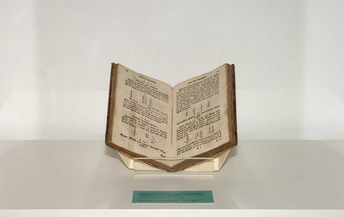 Aufgeschlagen in einer Vitrine steht Jakob Friedrich Maler's Schulbuch. Auf der Doppelseite werden mathematische Brüche behandelt.