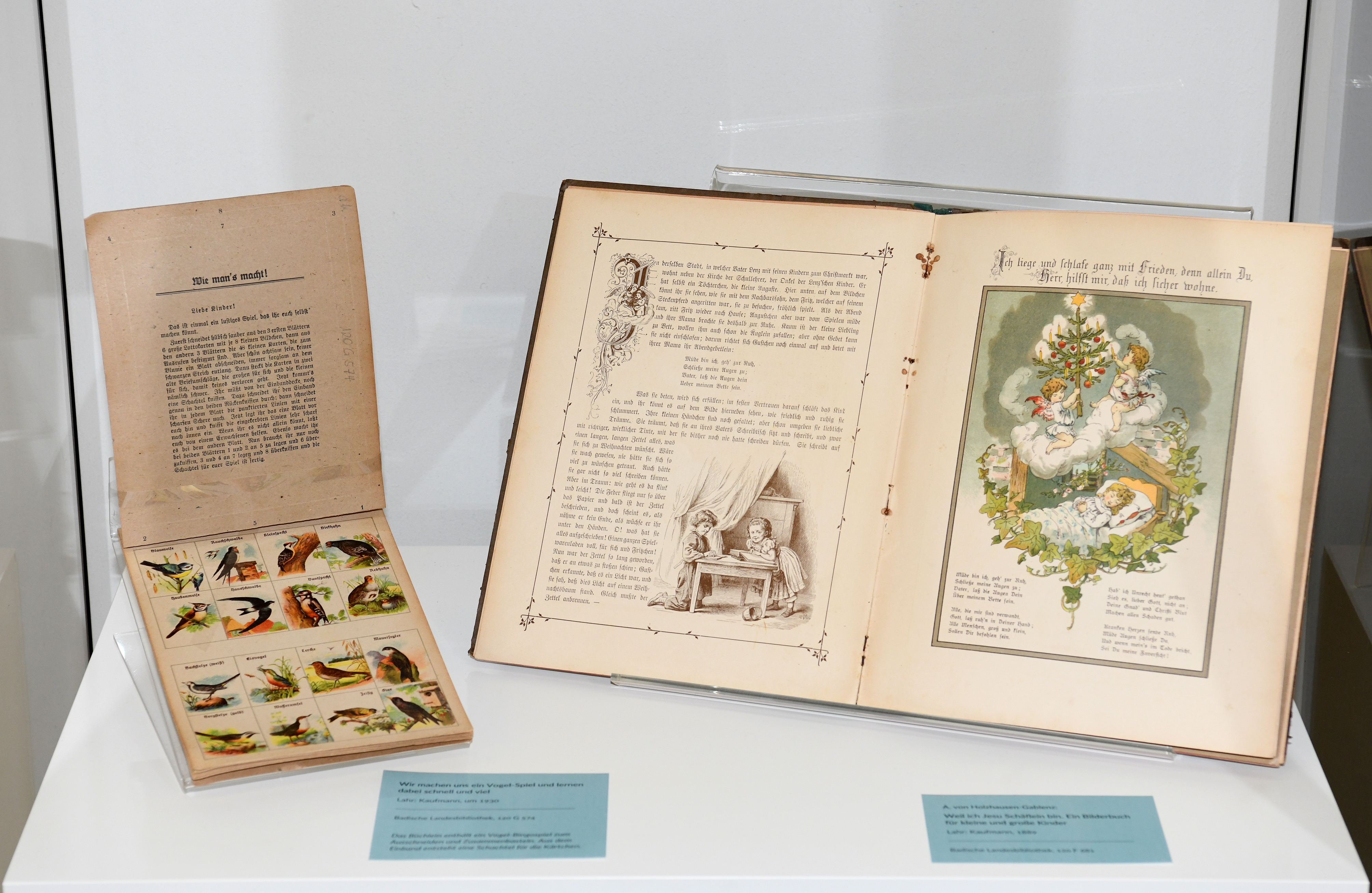 Ausgestellt sind zwei Künderbücher. Links ist eine Seite mit Vogel-Spiel. Rechts eine Doppelseite aus einem illustrierten Kinderbuch mit kleinen Engeln die einen Christbaum schmücken.