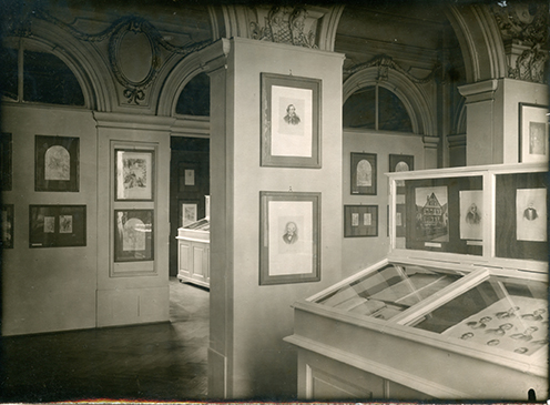 Schwarz-weiß Fotografie des ehemaligen Bibliothekssaal im Schloss. Blick auf die Schaukästen und gerahmten Fotografien und Zeichnungen an den Wänden.
