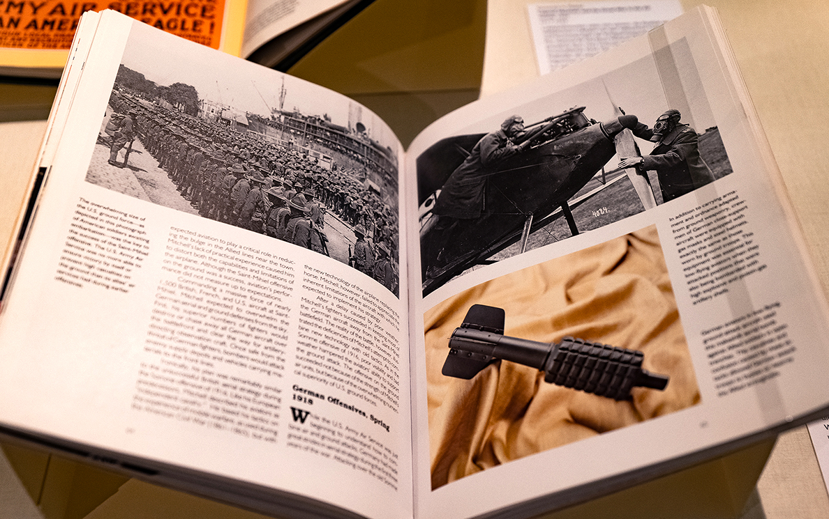 Geöffnetes Buch; links: Schwarz-Weiß-Foto einer Truppe der US-Army; rechts: Schwarz-Weiß-Foto zwei Kampfpiloten beim Flugzeug-Check; darunter: Farbfoto einer Fliegerbombe
