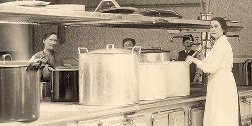 Schwarz-weiß Fotografie, die zwei Frauen dabei zeigt, wie sie in einer Kantinen-Küche in großen Töpfen kochen.