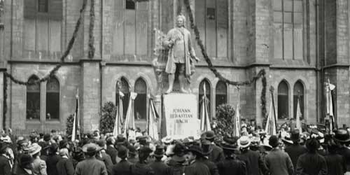 Das Bild zeigt die Einweihung eines Denkmals. Johann Sebastian Bach thront als übergroße Steinskulptur auf einem Sockel. Drumherum sind viele Menschen sowie Fahnen zu sehen, im Hintergrund die Kirche.
