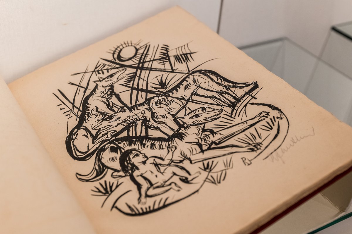 Zu sehen ist die aufgeklappte Seite eines Buches, auf welcher eine Illustration in schwarzer Tinte zu finden ist. Die Illustration zeigt drei ausgewachsene Wölfe mit einem menschlichen Baby, umgeben von Dschungelpflanzen.