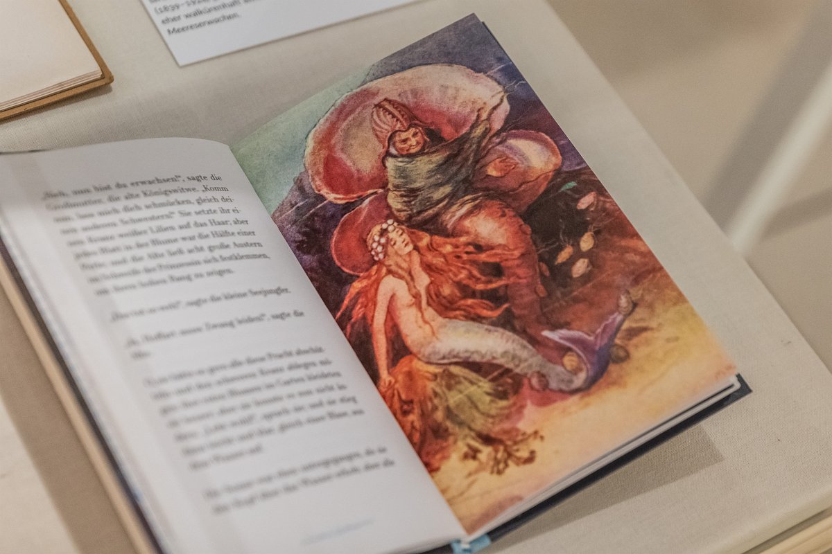 Eine Fotografie des Buches „Die kleine Meerjungfrau“. Der Fokus liegt auf einer Illustration der kleinen Meerjungfrau: Diese sitzt auf einer Koralle, neben ihr tritt die böse Meereshexe aus einer großen Muschel hervor.